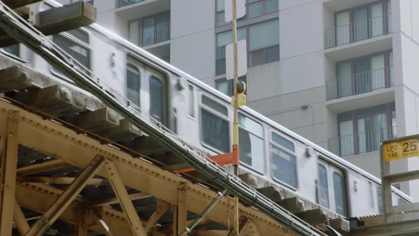 A subway train travels on an elevated track in a city with an apartment building in the background