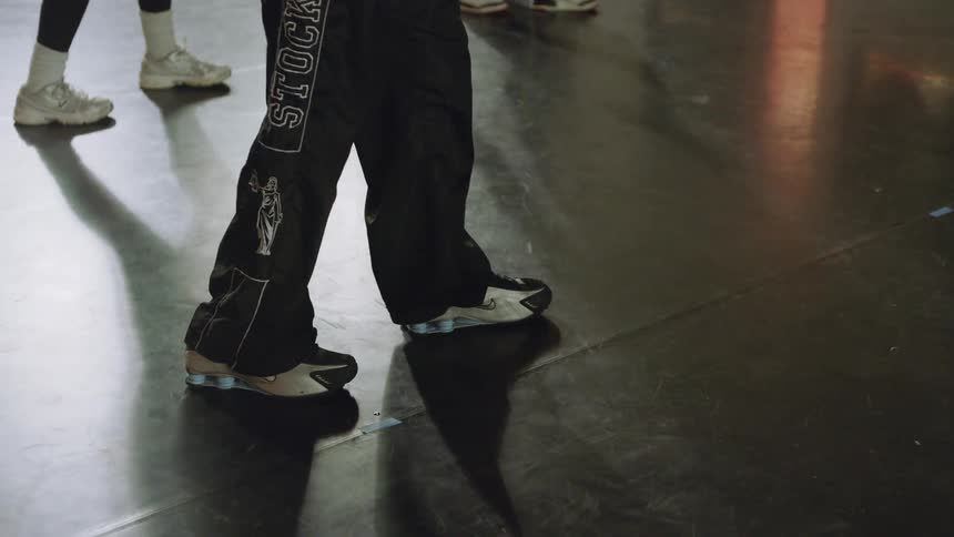 A close-up of a dancer's feet in sneakers performing rhythmic footwork on a dark, reflective studio floor