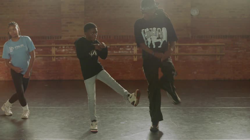 A male hip-hop instructor teaches a young boy a dance routine in a sunlit studio while another student practices nearby