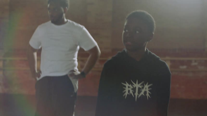 A young Black boy practices intricate dance footwork in slow motion in a sunlit studio as his coach watches from the background