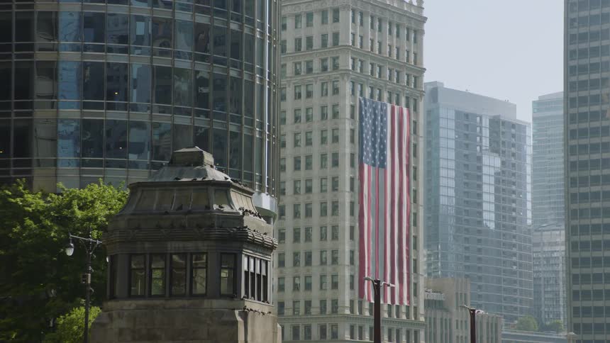 A massive American flag is displayed on a building, framed by a mix of modern glass skyscrapers and historic city architecture