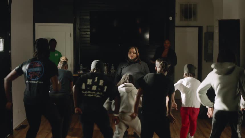 An instructor leads a group of young dancers through a synchronized hip-hop routine in a dimly lit studio