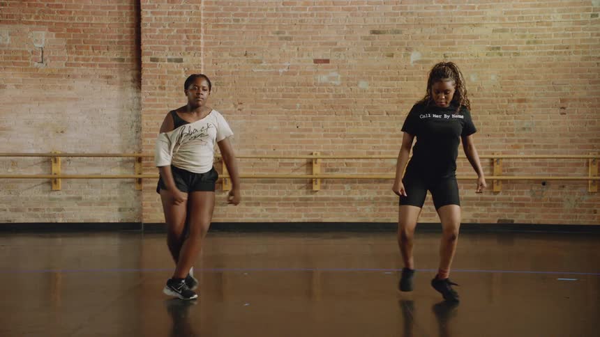 Two female dancers perform a synchronized hip-hop routine in slow motion inside a dance studio