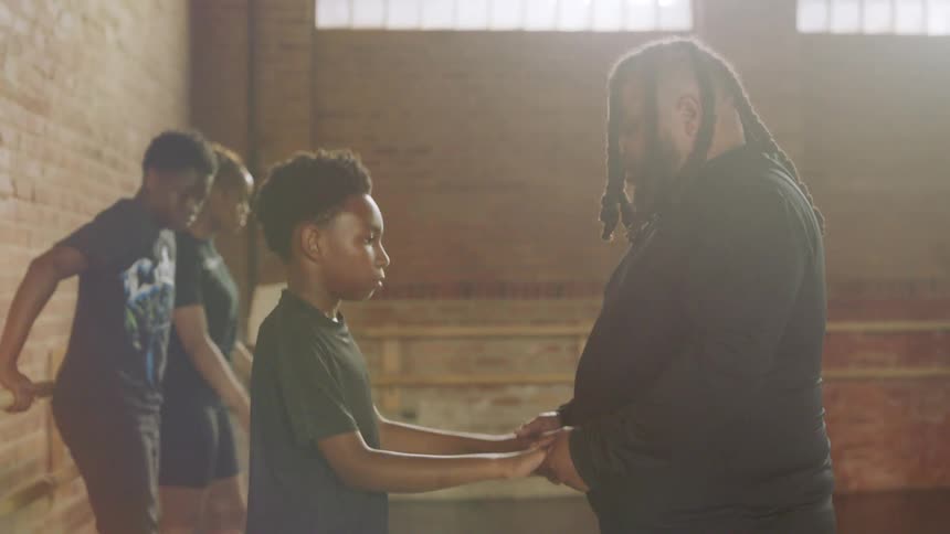 A dance instructor with dreadlocks patiently teaches a young boy footwork in a sunlit studio