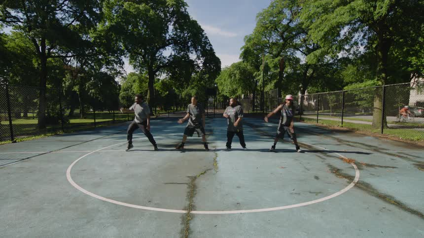 Four talented men perform an energetic and synchronized hip-hop dance routine in the center circle of a park basketball court