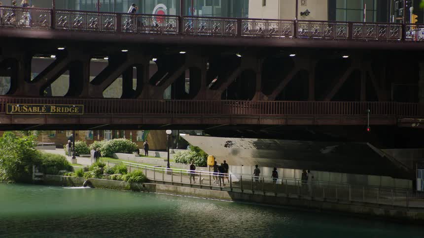 Pedestrians walk along the Chicago Riverwalk under the historic DuSable Bridge on a sunny day