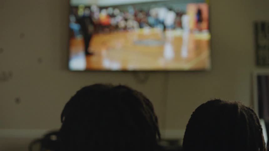 Backs of two people's heads as they watch a dance competition on a large television mounted on the wall