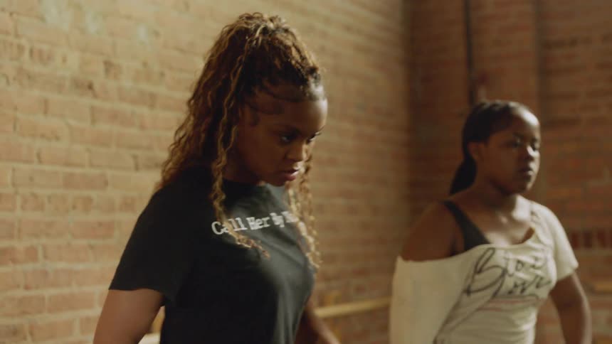 A group of young dancers practices a hip-hop routine in slow motion inside a sunlit studio with brick walls