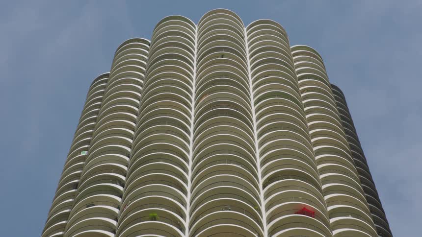 A time-lapse from a low angle shows a modern cylindrical skyscraper with repeating balconies as clouds pass by in the blue sky