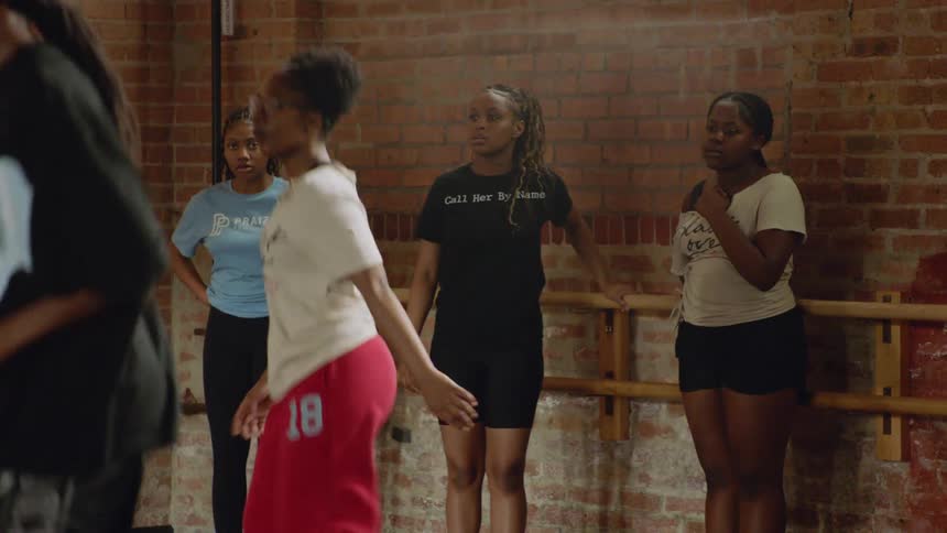 An energetic female instructor leads a hip-hop dance class, teaching a new routine to a group of young women in a brick-walled studio