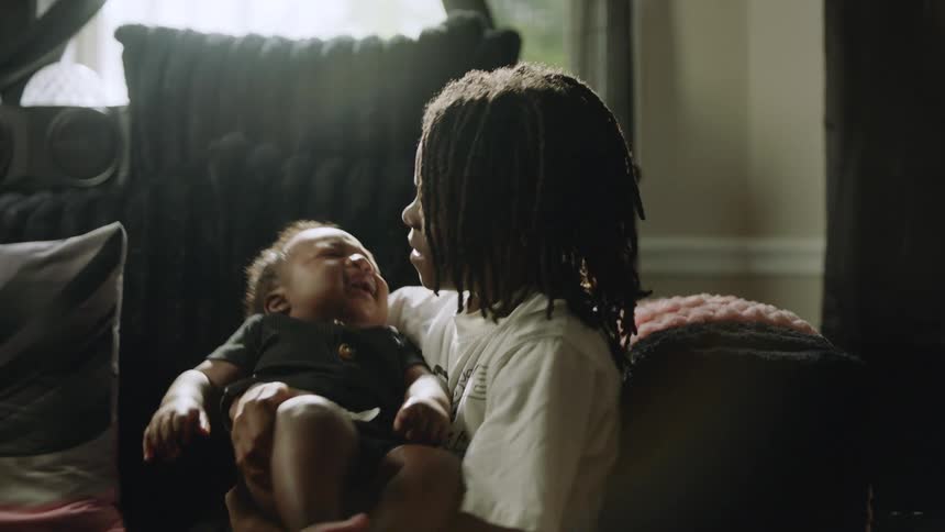 A caring older sibling with dreadlocks gently holds their crying baby sibling, trying to soothe them in a softly lit room