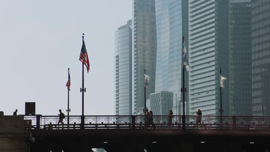 Silhouettes of people walking and jogging across a city bridge with American flags waving against a backdrop of modern skyscrapers