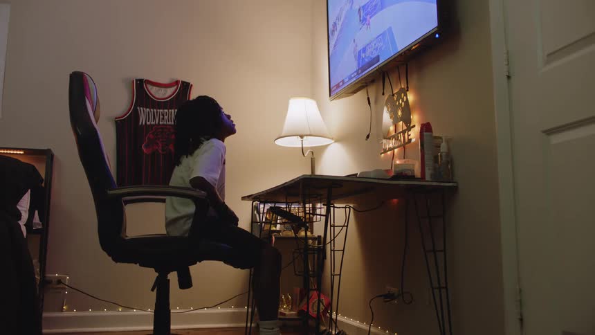 A young person sits in a gaming chair in their dimly lit bedroom, intently focused on a basketball video game on their television