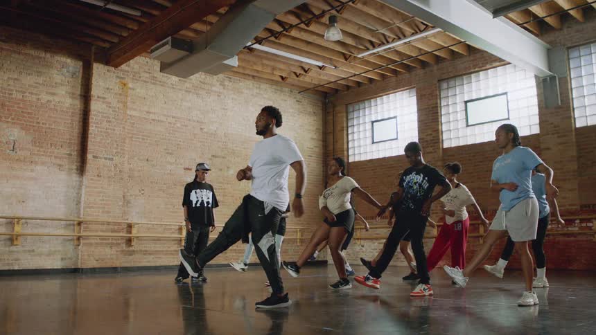 A male instructor leads a diverse group of teenagers in a hip-hop dance class inside a spacious studio in slow motion