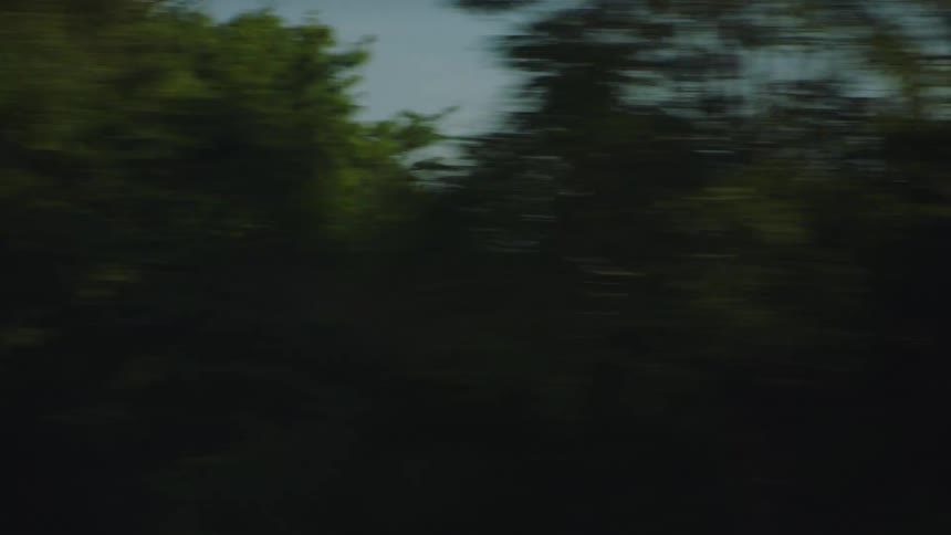 A blurry point-of-view shot from a moving vehicle captures the fast-paced motion of passing under bridges and by cityscapes and suburban landscapes