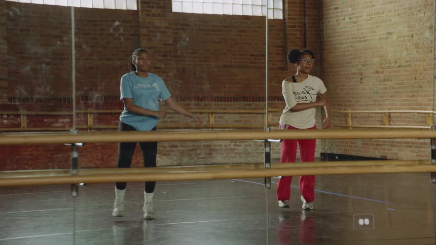 Two young women happily practice a synchronized dance routine in slow motion in a beautiful studio with a brick wall