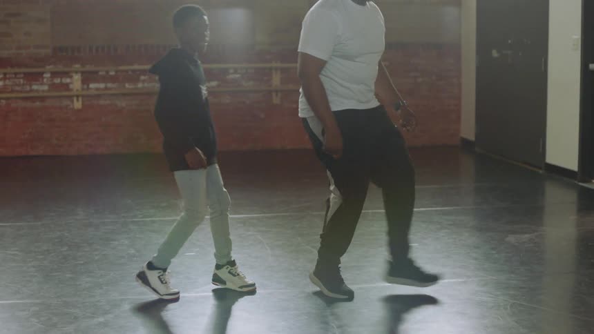 A dance teacher shows a young boy some cool footwork in a sunlit studio as they practice together