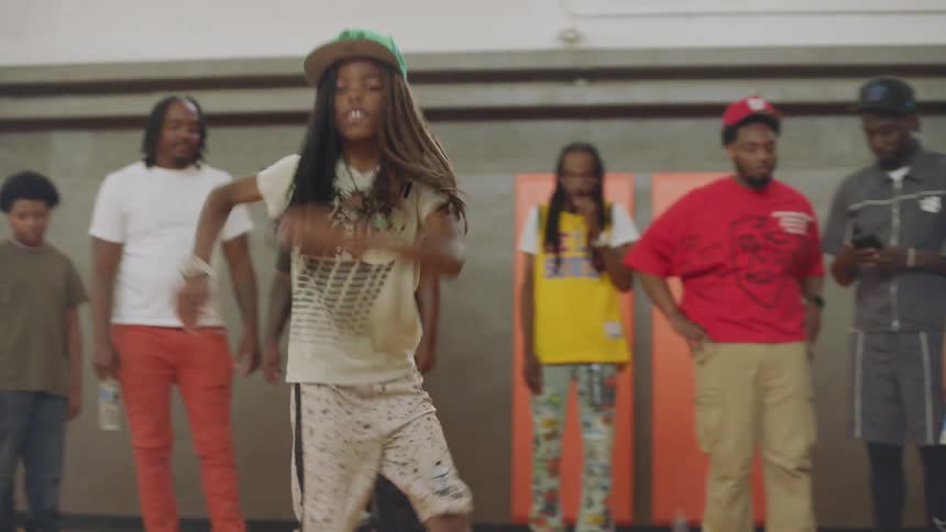 A confident young dancer with long dreadlocks shows off their impressive hip-hop moves in a gym while a crowd watches