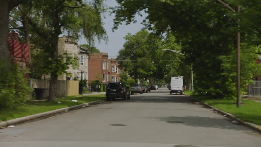 A driving plate shot through a rundown urban neighborhood, showing a local market, an alleyway with dumpsters, and vacant lots next to residential homes
