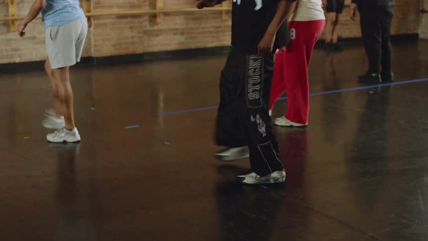 A dance instructor leads a group of dancers in a hip-hop class inside a studio, with the camera tilting down to show their intricate footwork in slow motion