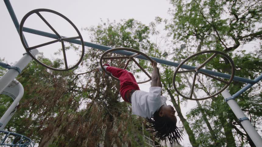 From a low angle, a happy young boy with dreadlocks hangs upside down from a jungle gym in slow motion