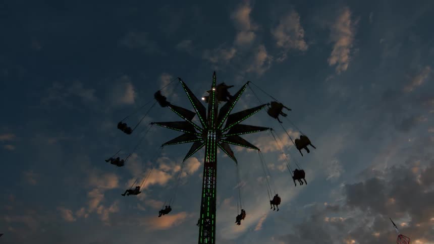 Silhouetted figures on a lighted carnival swing ride spin against a dramatic evening sky with sunset-tinted clouds