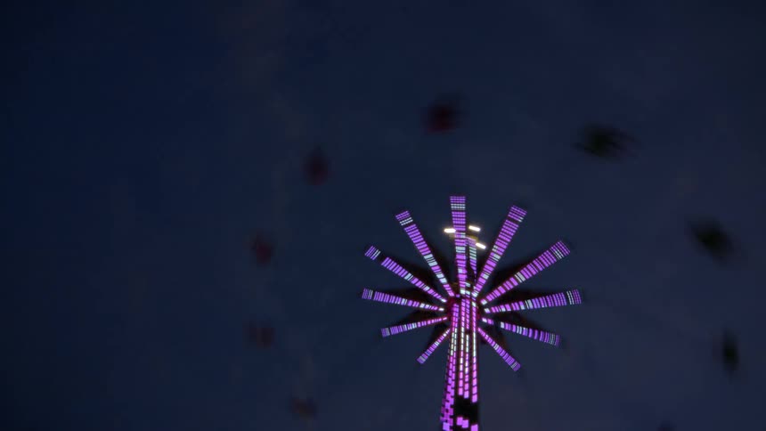 A low-angle view of a spinning carnival swing ride illuminated by vibrant green and purple LED lights against the dark night sky