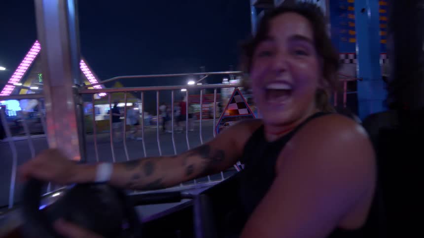 A joyful woman with tattoos laughs hysterically while riding the bumper cars at a brightly lit amusement park at night