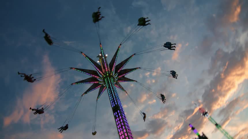 A rotating low-angle view of a brightly lit swing ride spinning people against a beautiful sunset sky at a carnival