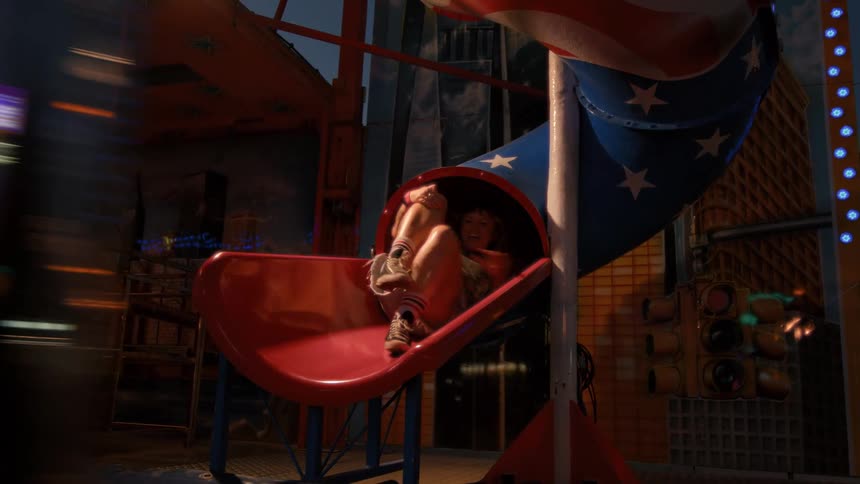 A joyful woman with a mullet and tattoos slides down a funfair slide at night, laughing as she reaches the bottom