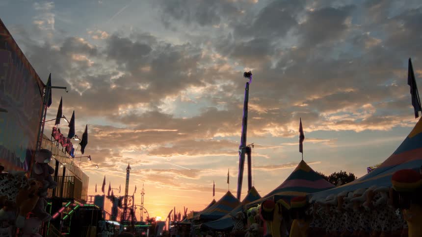 A bustling carnival with vibrant rides and game booths is silhouetted against a beautiful and dramatic sunset sky