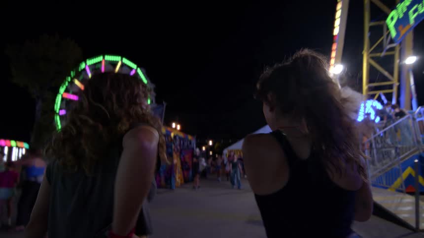 Two friends walk through a vibrant, neon-lit carnival at night in slow motion, heading towards the rides and games