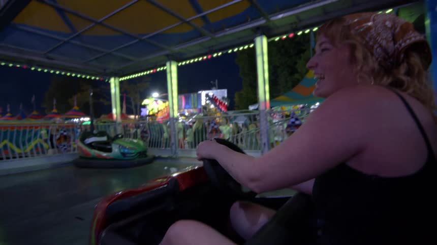 Two women laugh and smile while having fun riding the brightly lit bumper cars at a lively carnival at night