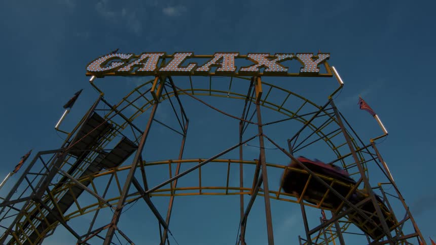 A low angle view shows cars speeding by on the 'Galaxy' roller coaster at a carnival under a blue sky