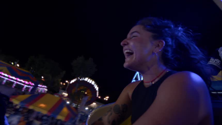 In slow motion, a joyful young woman with tattoos laughs and screams while on a spinning carnival ride at night with friends