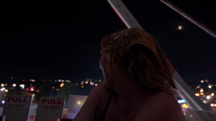 A young woman with a bandana smiles and enjoys the view from a Ferris wheel overlooking a brightly lit carnival at night