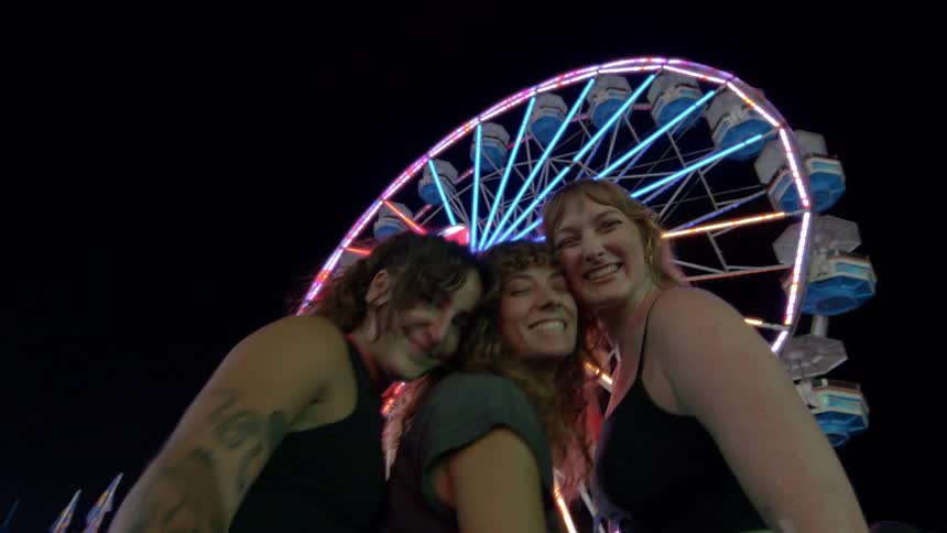 A low-angle selfie view of three happy friends posing and laughing together at a nighttime carnival with a colorful Ferris wheel in the background