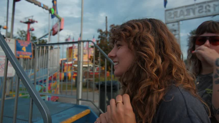 A young woman with curly hair smiles with excitement while watching a ride at a local carnival