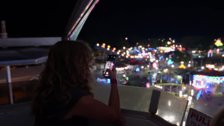 A young woman on a Ferris wheel at night takes photos of the vibrant carnival lights below with her smartphone before looking out in amazement