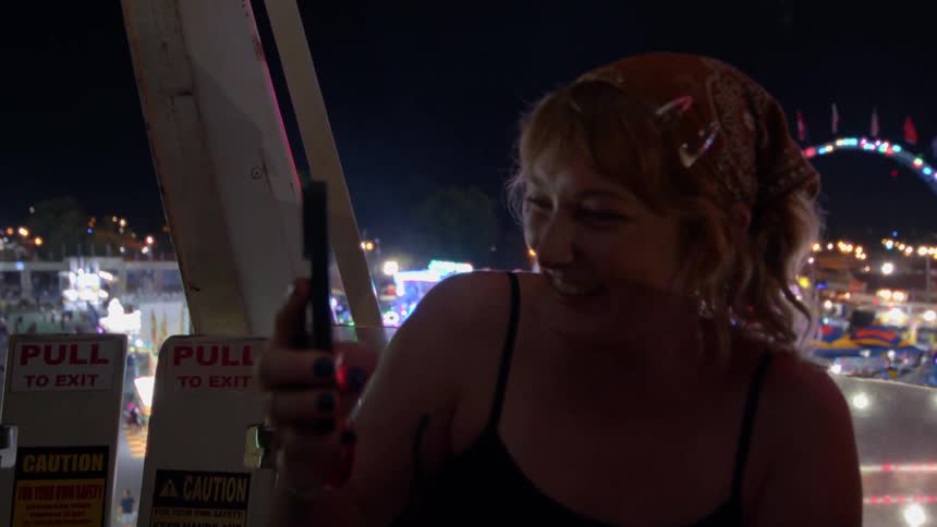 A joyful woman captures a moment on her phone while riding a Ferris wheel high above a bustling night carnival