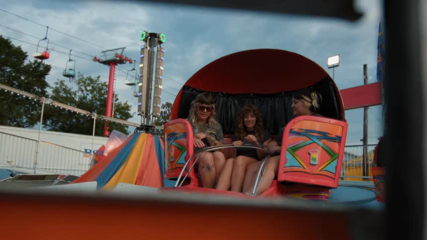 Three young female friends laugh and talk together on a spinning ride at a carnival in the evening