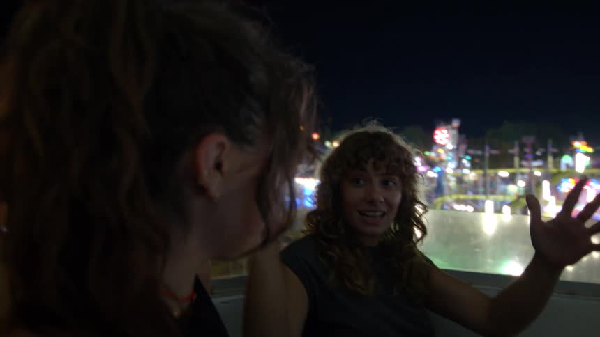 An animated young woman excitedly talks with her friend while riding a Ferris wheel with the colorful carnival lights in the background