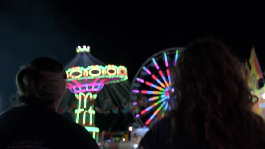 From behind, a woman with long hair watches the colorful, spinning lights of a Ferris wheel and chair swing ride at a carnival