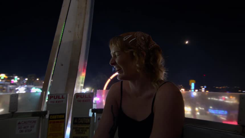 A happy young woman laughs in slow motion while enjoying the view from a Ferris wheel at a vibrant carnival at night