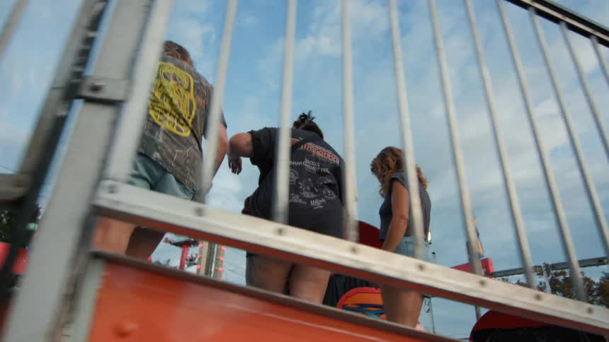 A low-angle, slow-motion view shows three friends getting onto a spinning carnival ride from behind a metal gate