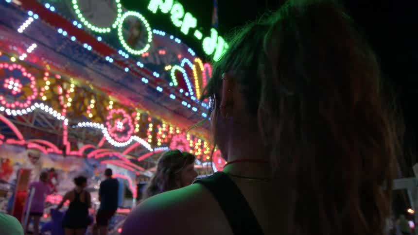 Over the shoulder view of a woman with a ponytail watching a brightly lit, flashing carnival ride at night