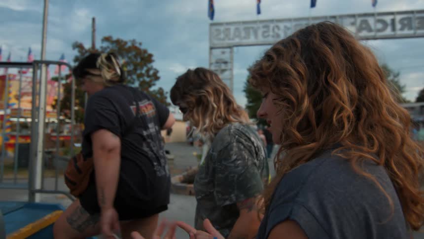 A group of young women with long hair walk up the stairs to board a carnival ride at a local fair in slow motion