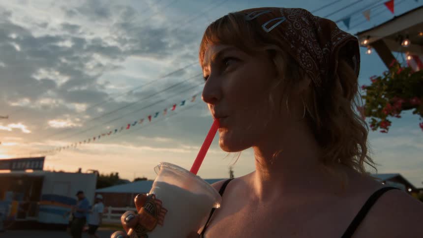 A smiling young woman enjoys a milkshake in slow motion at an outdoor fair during a beautiful sunset