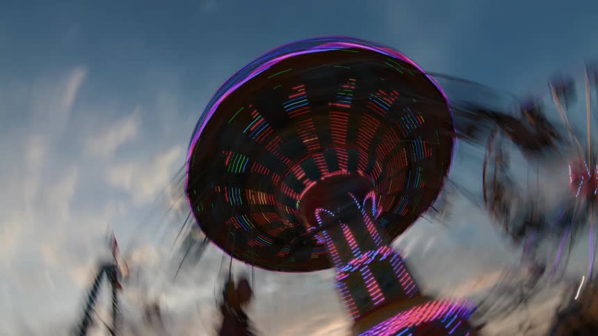 A brightly lit carnival swing ride spins into a colorful motion blur against the evening sky