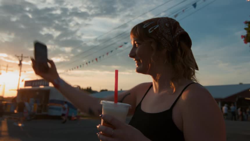 A happy young woman with a bandana and a drink takes a selfie with her smartphone at a summer fair during sunset
